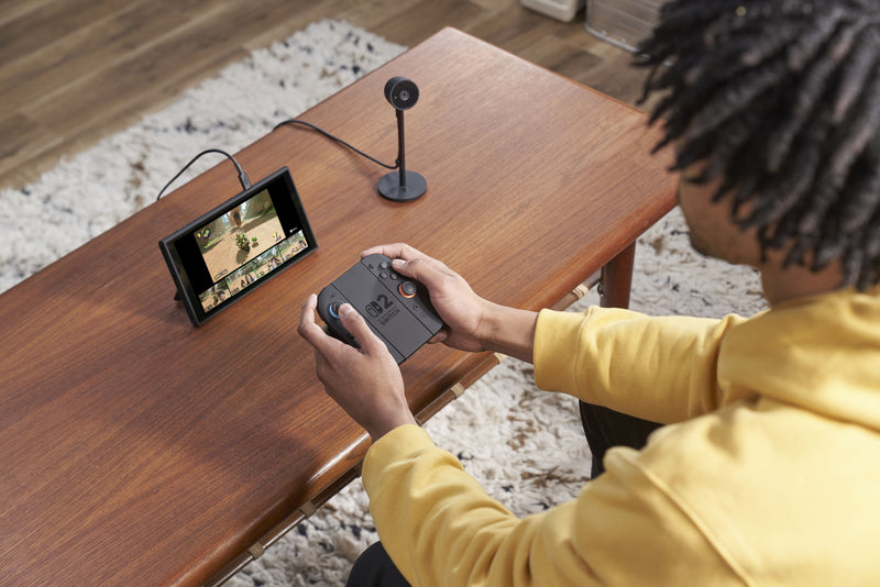 Person using a Nintendo Switch 2 gaming console with a screen on a wooden table